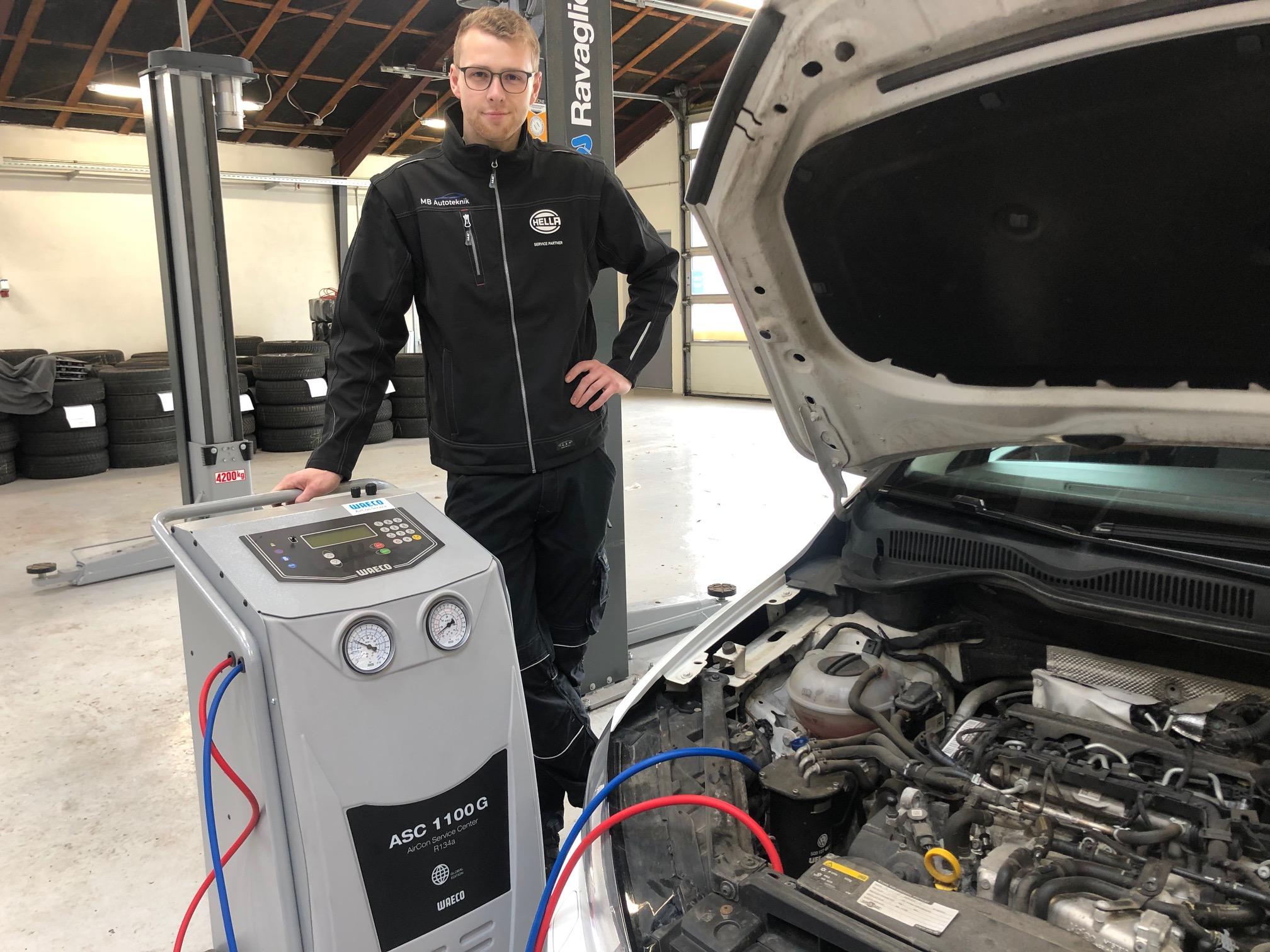 En mekaniker står ved en airconditionmaskine i et bilværksted med en åben motorhjelm.