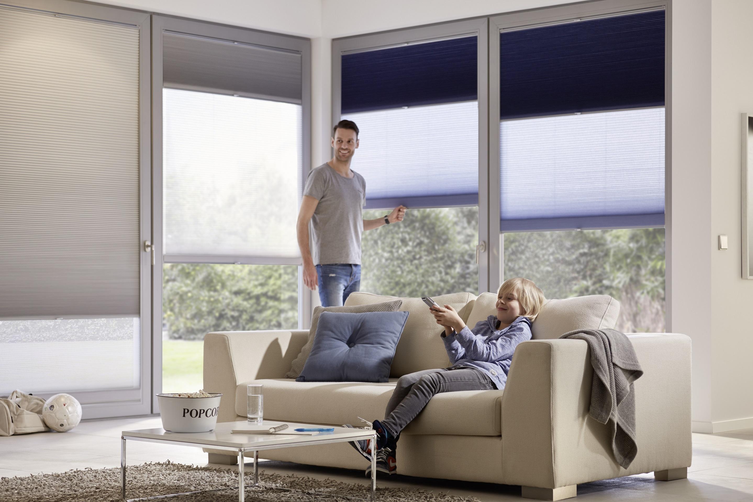 En mand og en kvinde sidder i en stue med store vinduer og moderne persienner, mens de slapper af på en lys sofa.