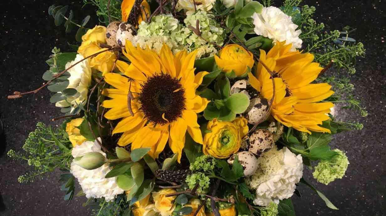 En farverig buket med solsikker, ranunkler og hvide blomster, prydet med grønne blade og grene.