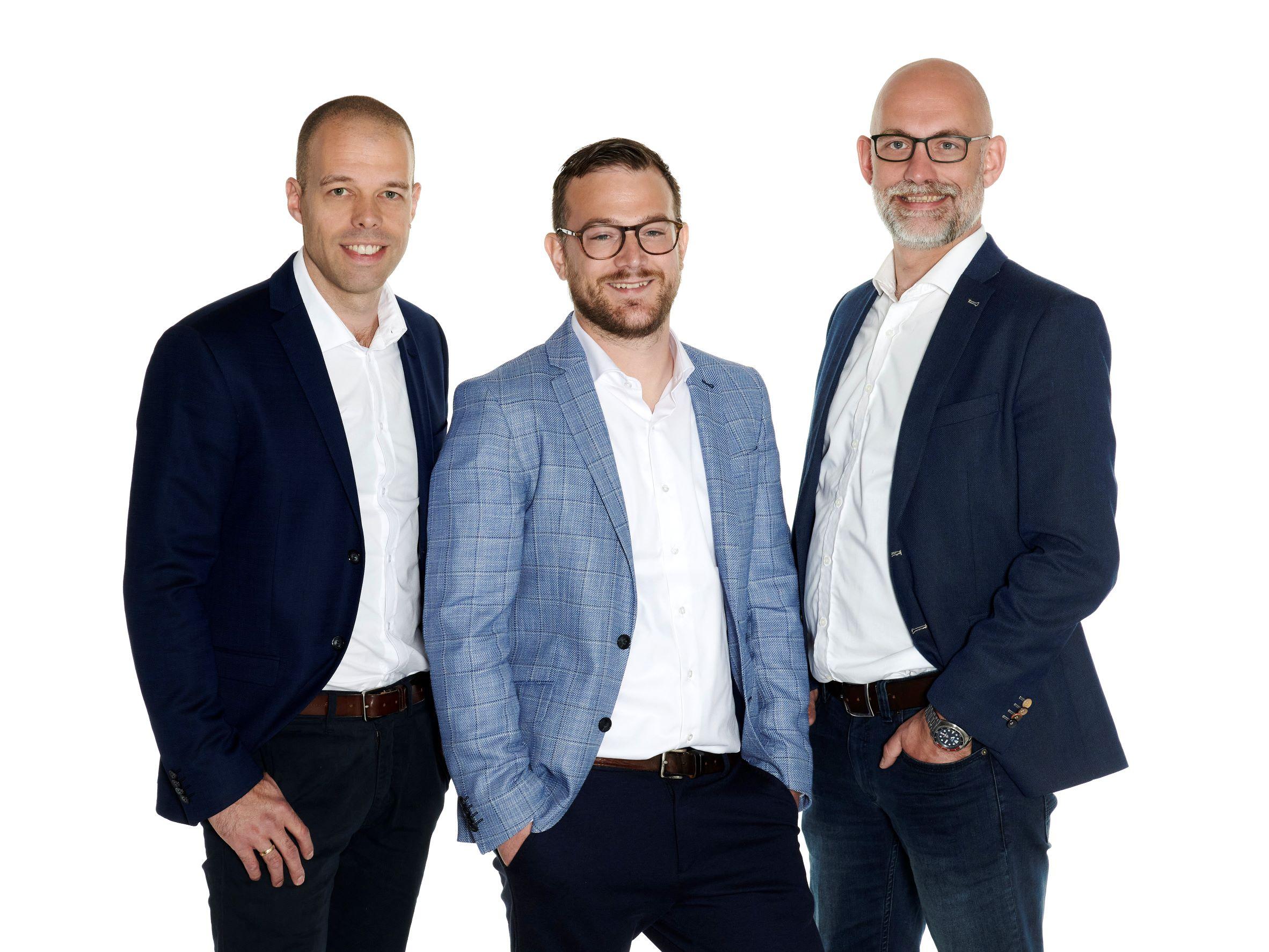 Tre mænd står sammen i formelle jakkesæt med smilende ansigtstræk på hvid baggrund.