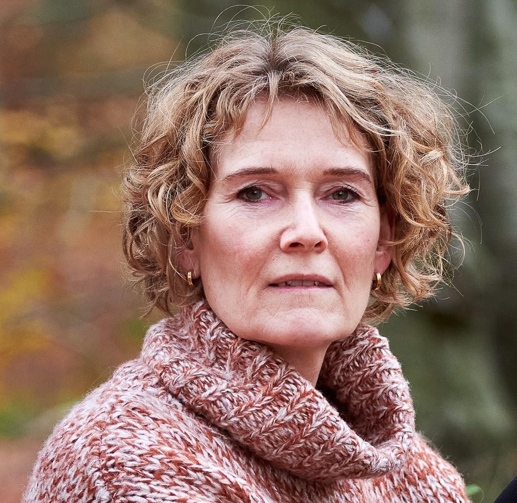 En kvinde med krøllet hår iført en strikket sweater står i en skov med efterårsfarver.