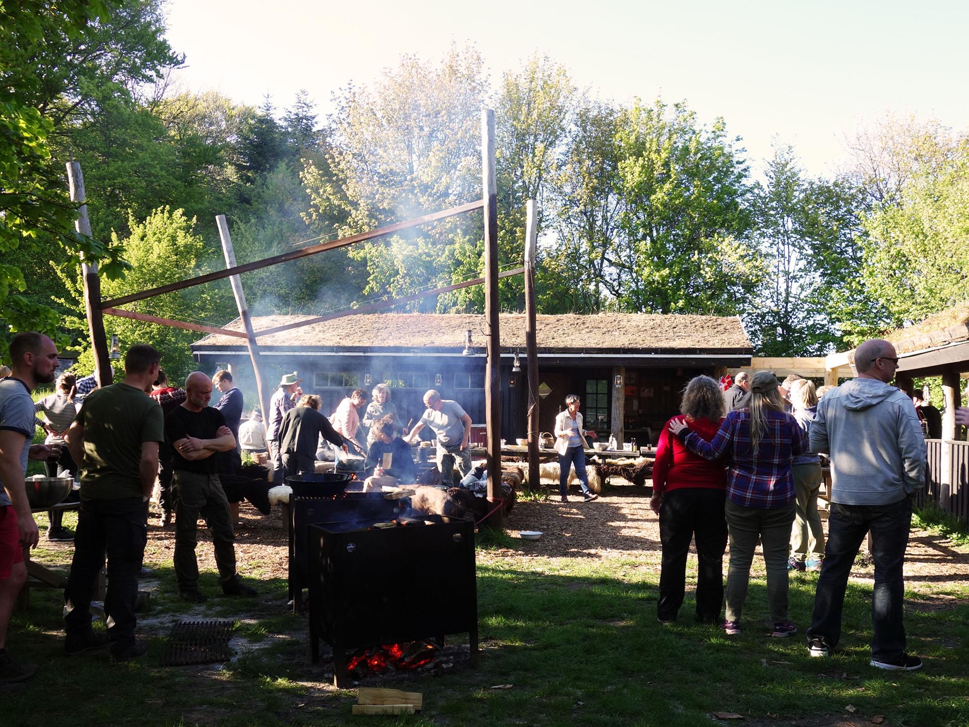 Mennesker samles omkring en grill i en skov, mens røg stiger op fra bålet og en hytte ses i baggrunden.