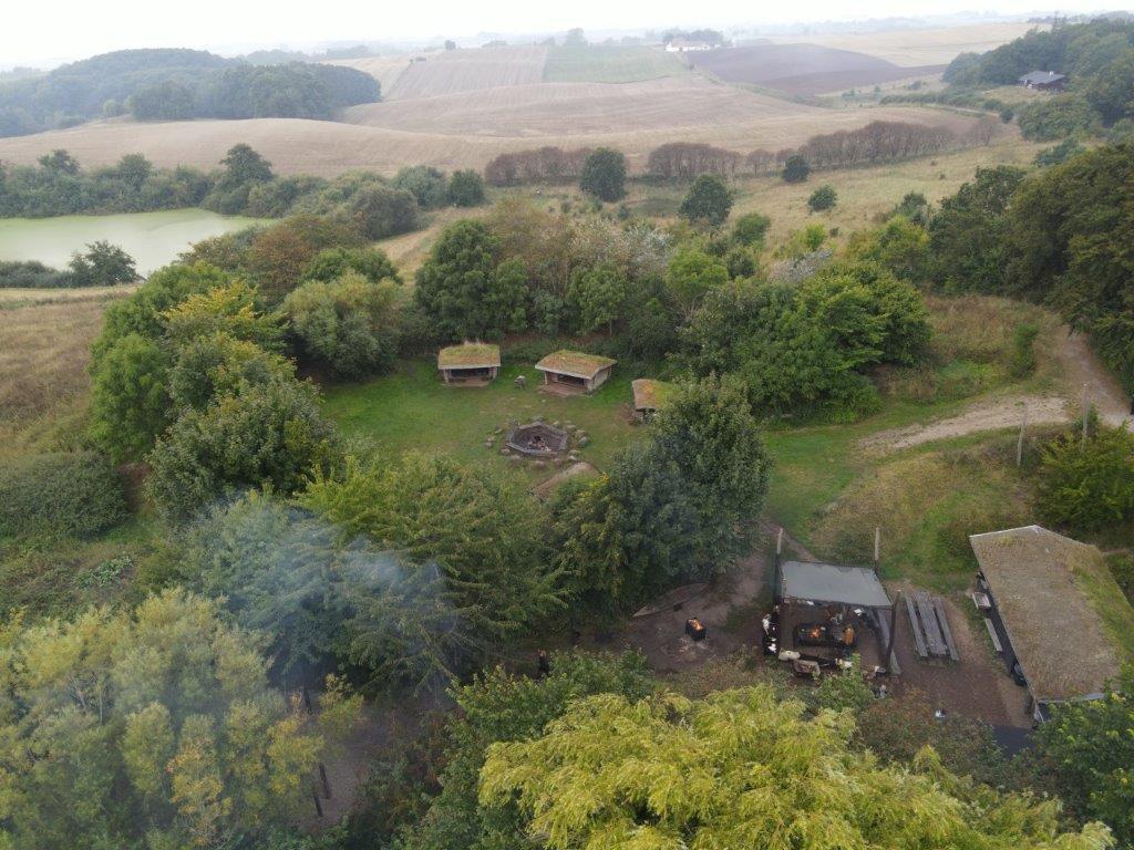 Luftfoto af en grøn græsplæne med bålplads og shelter i et naturskønt landskab.