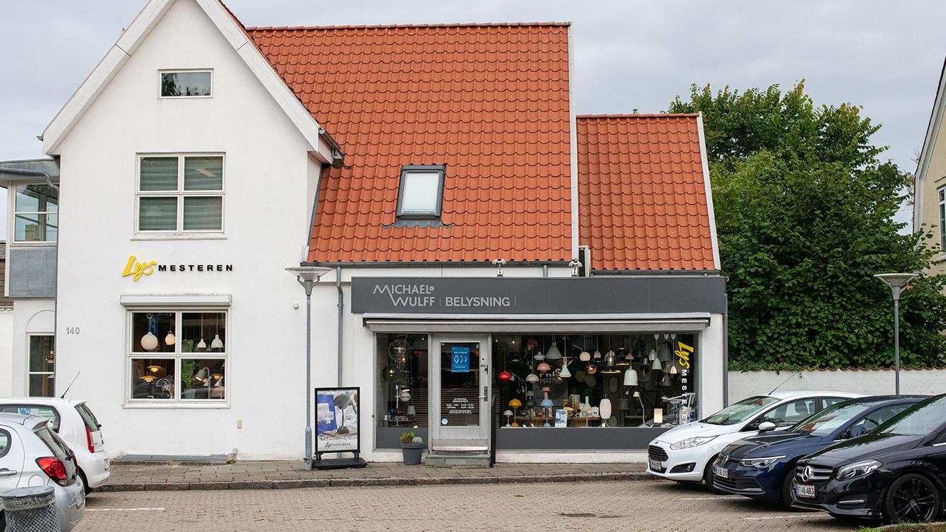 En butik med grå facade og rødt tag, fyldt med lamper, beliggende i en by med parkerede biler foran.