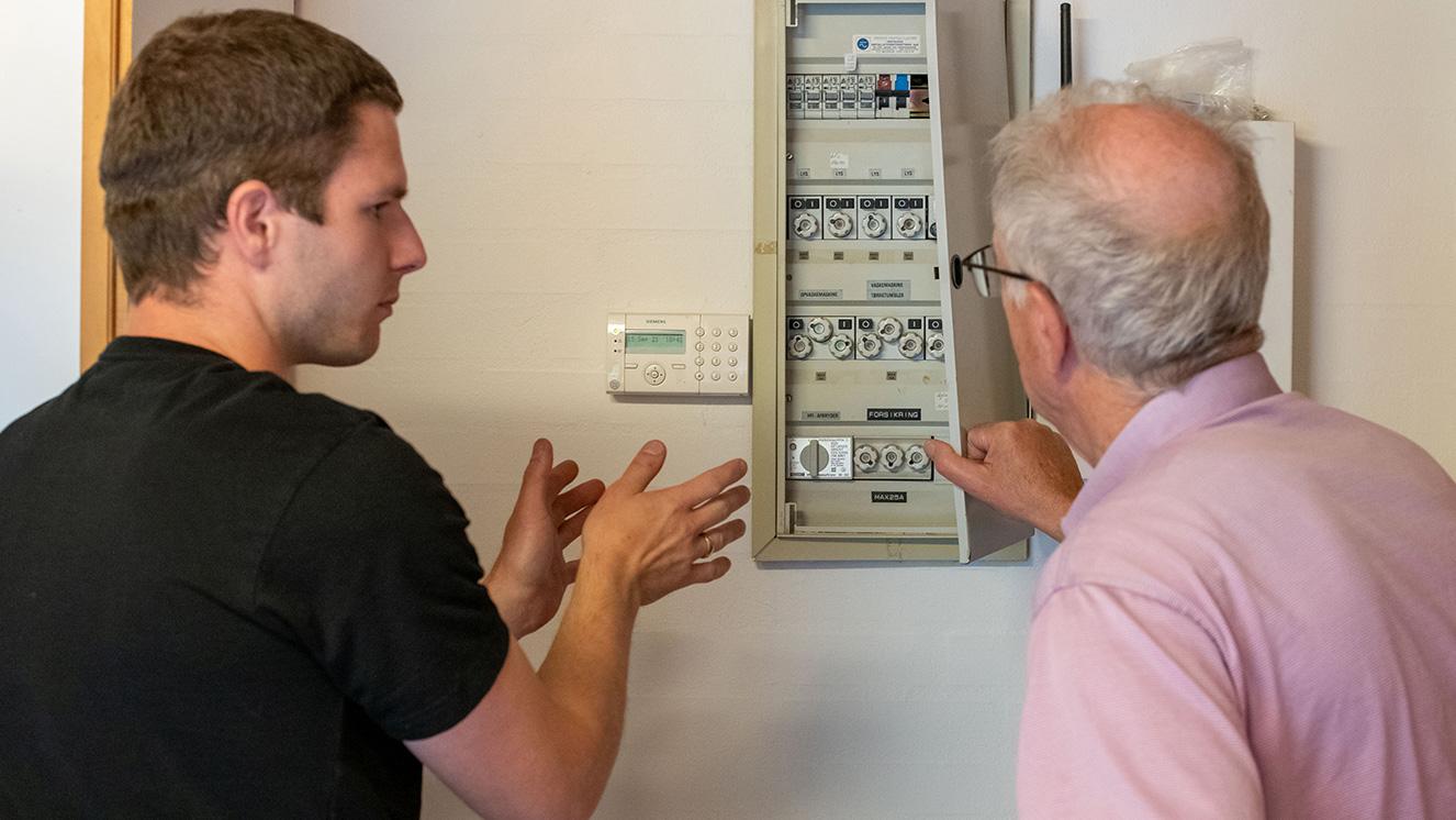 To mænd, der diskuterer en elektrisk tavle i et hjem, mens de peger på forskellige komponenter.