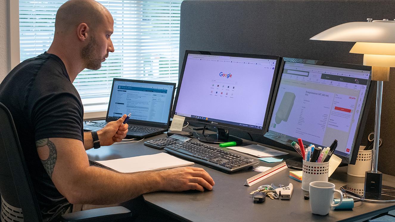 Mand arbejder ved et skrivebord med to computere, noter og kontorartikler omkring sig.