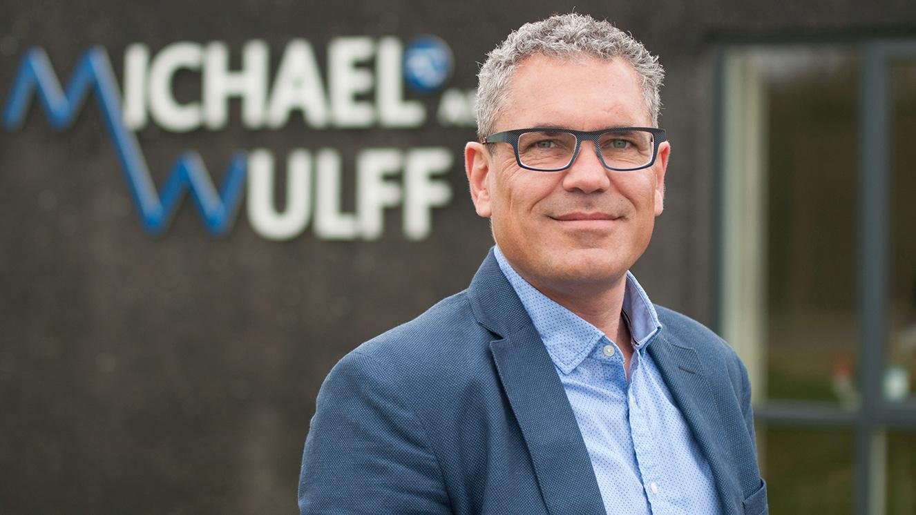 En mand med briller står foran en bygning med navnet "Michael og Wulff" synligt i baggrunden.