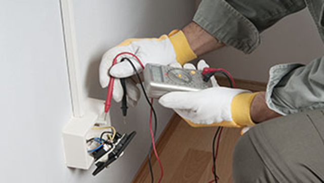 En person i handsker måler elektrisk spænding med et multimeter ved en stikkontakt på væggen.