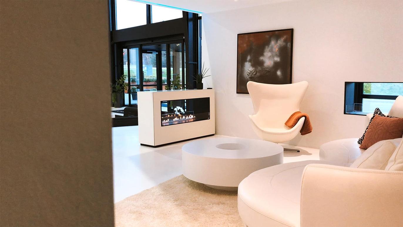 Moderne stue med hvid lænestol, rund sofabord og elegant pejs i et lyst rum med store vinduer.