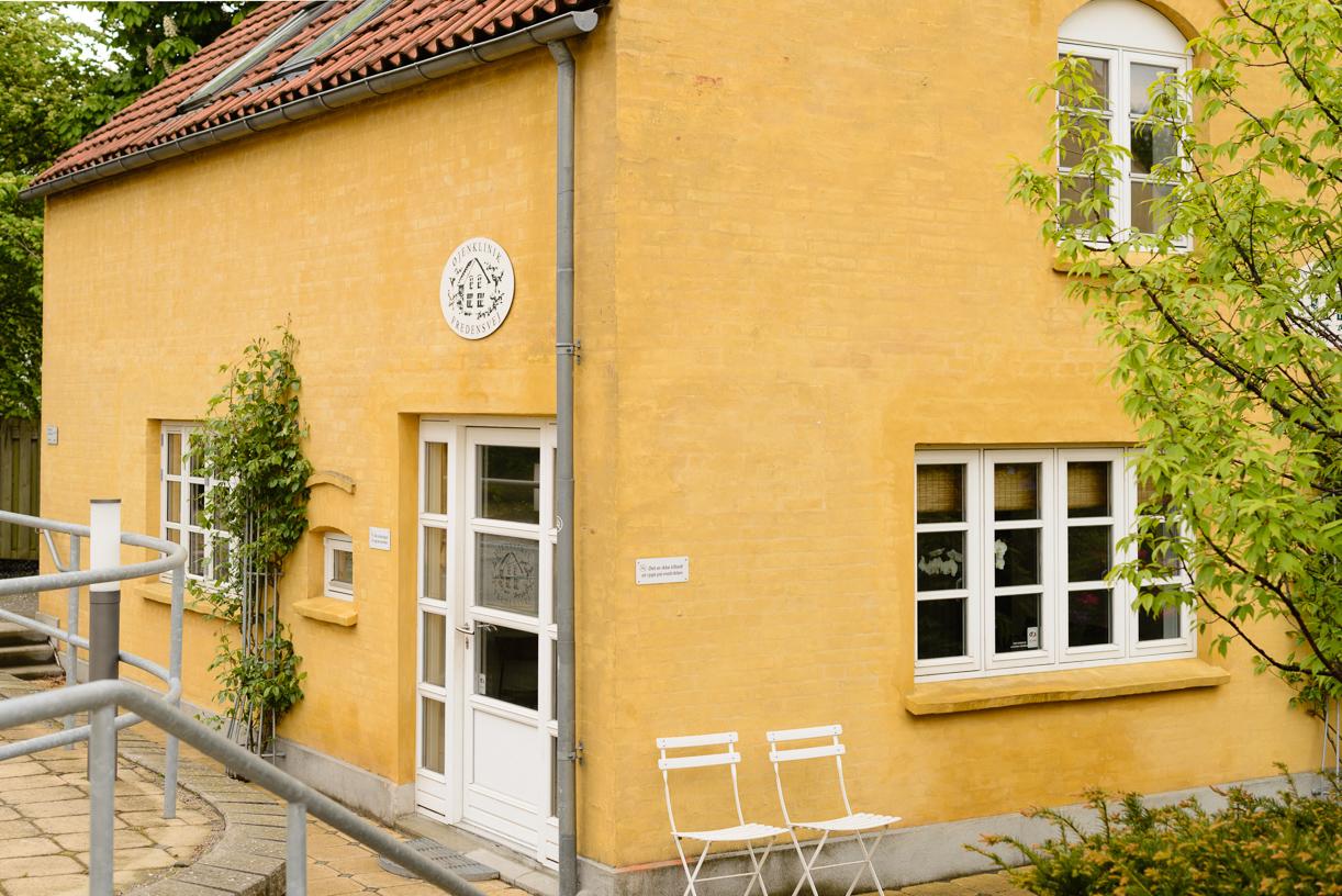 Gult hus med hvid dør og vinduer, omgivet af grønne planter og en lille terrasse med to stole.