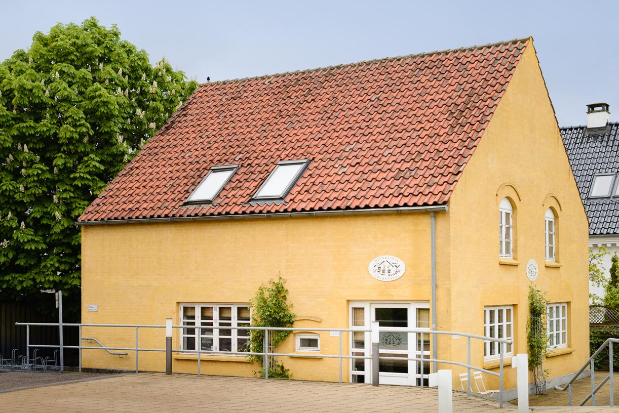 Gult hus med skråtag og vinduer, omgivet af grønne træer og en grusvej.
