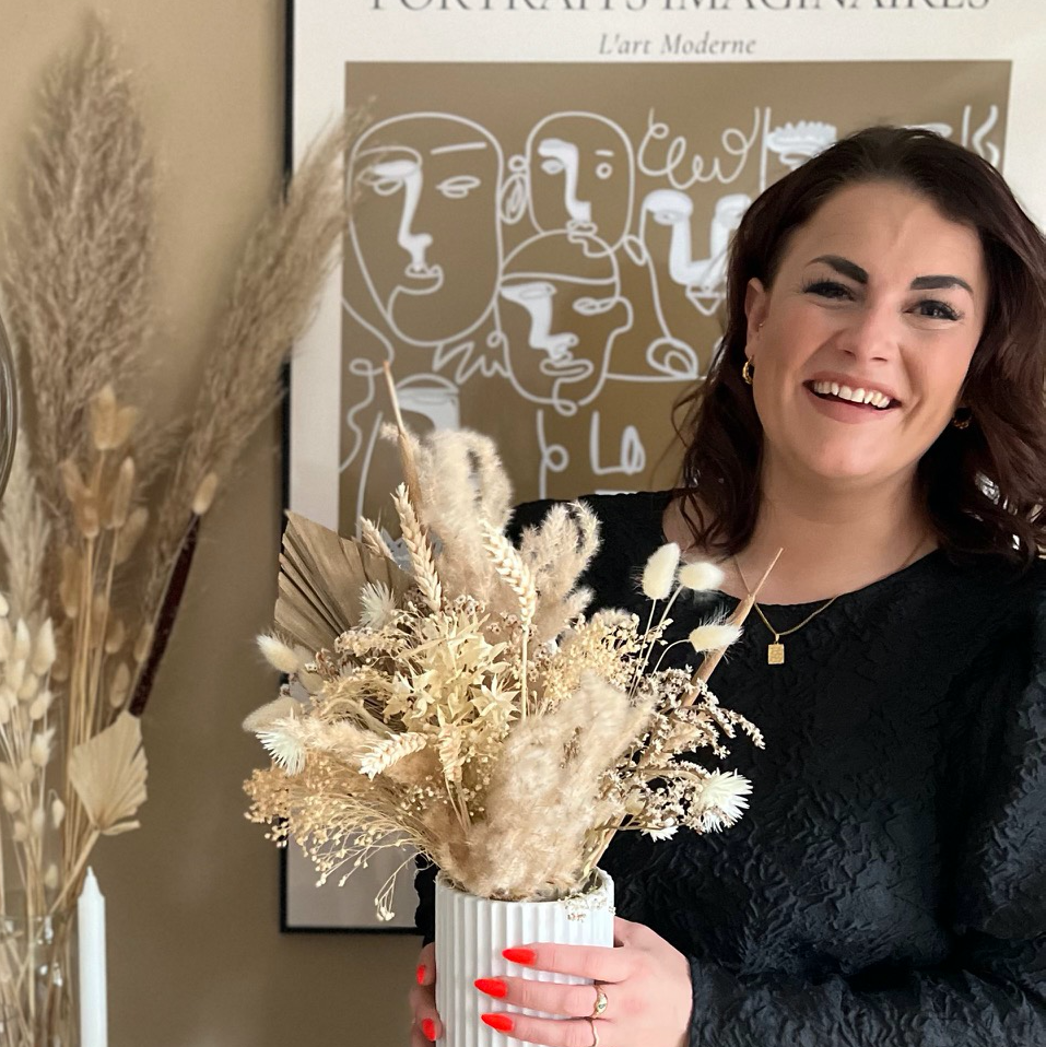Kvinde med brunt hår holder en vase med tørrede blomster foran en kunstplakat.