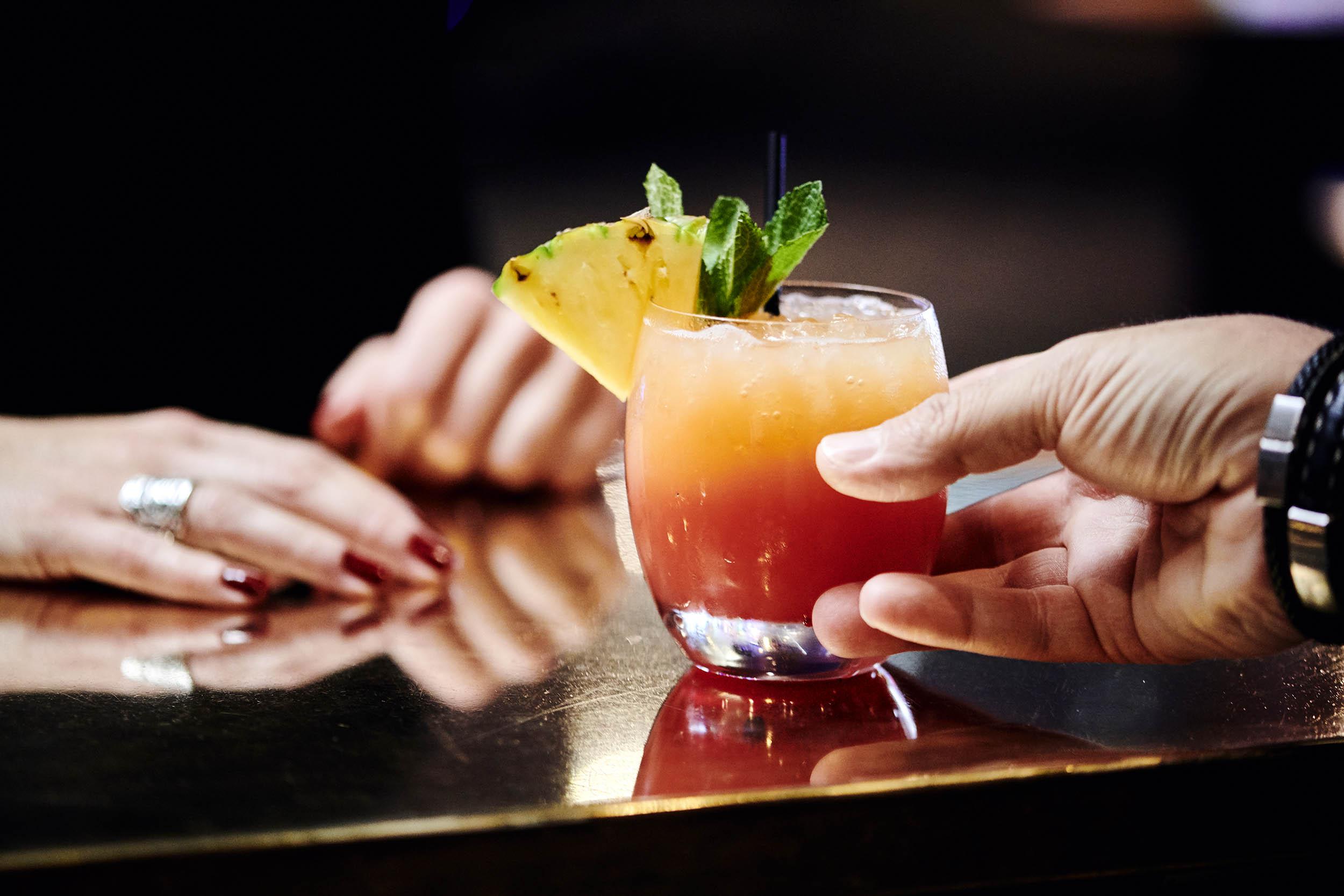 En farverig cocktail med ananas og mynte, der bliver rakt over en bar.