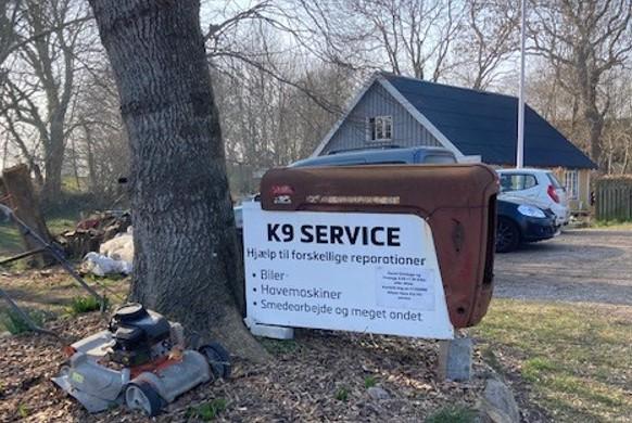 Skilt med teksten "K9 SERVICE" ved siden af en gammel bil og en græsslåmaskine under et træ.