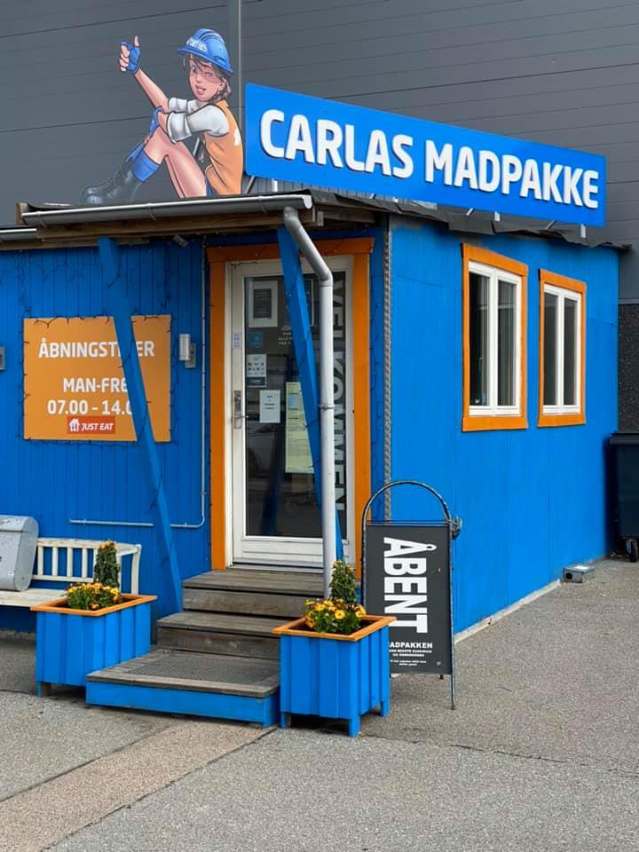 Blå madvogn med skiltet "Carla's Madpakke" og åbningstider, beliggende på en grå asfaltplads.