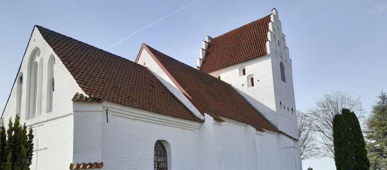 En hvid kirke med rødt tag og klokketårn, omgivet af træer under en klar blå himmel.