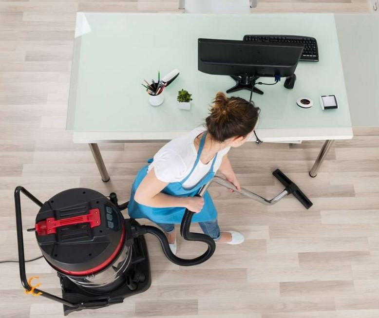 En kvinde støvsuger et kontor med en stor støvsuger, mens hun står ved et skrivebord med en computer.