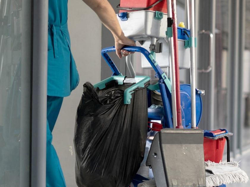 En person i arbejdstøj trækker en rengøringsvogn med en sort affaldssæk og rengøringsudstyr.