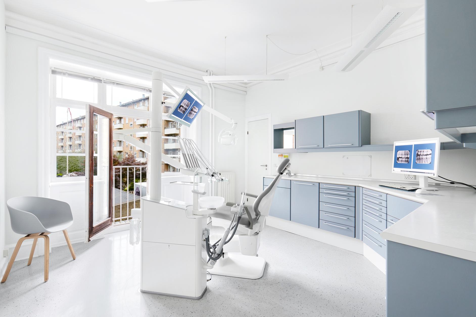 Moderne tandlægeklinik med lys indretning, tandlægestol og computere ved vinduet.