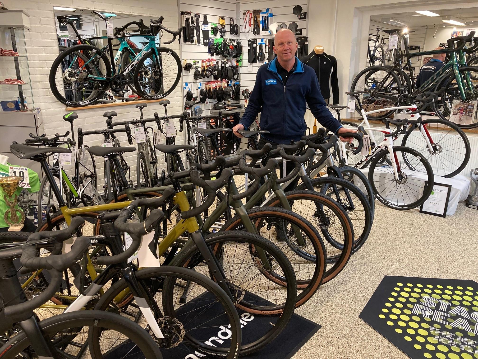 En mand står i en cykelbutik med flere cykler i forgrunden og cykeludstyr i baggrunden.