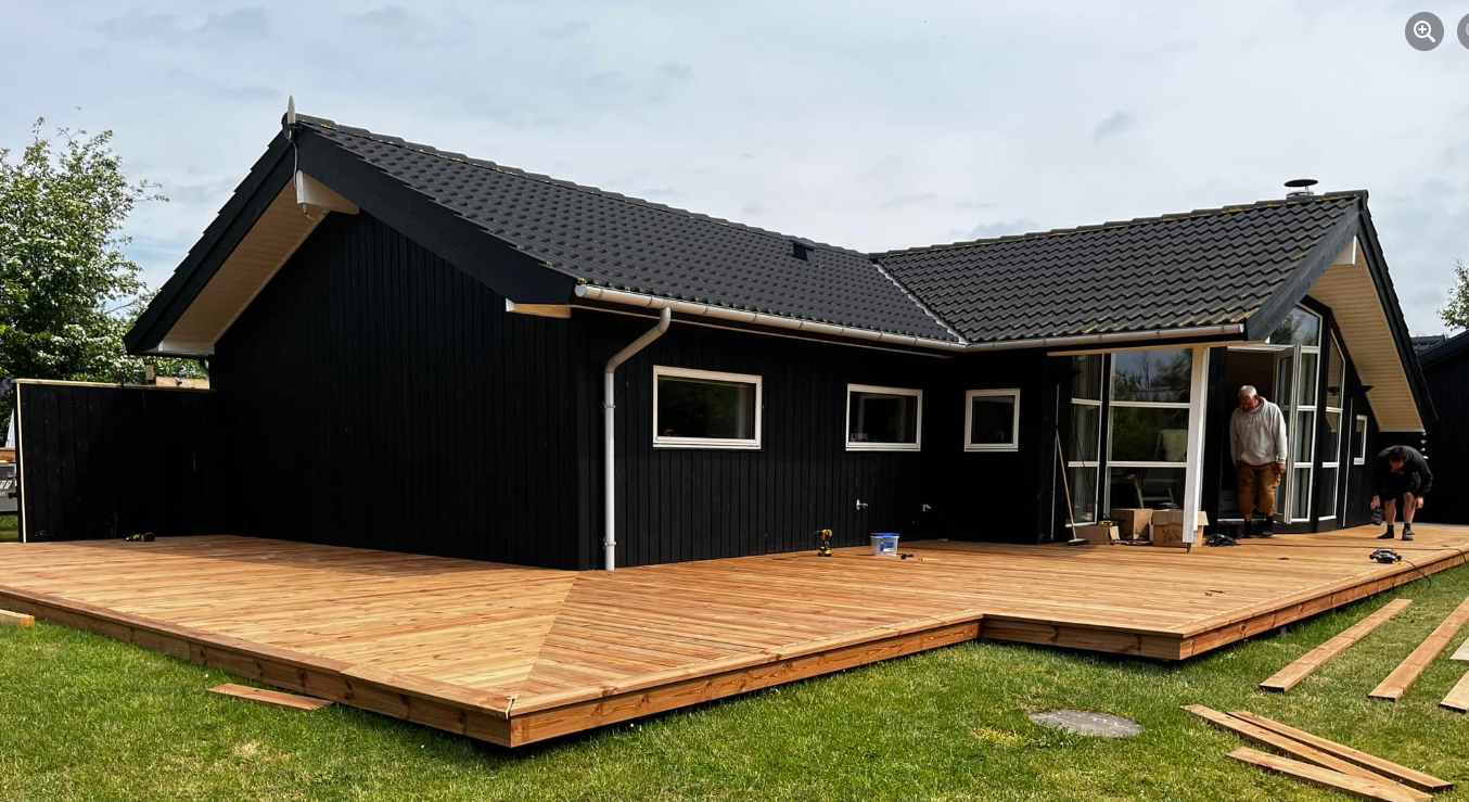 Moderne sort hus med træterrasse og store vinduer, omgivet af græsplæne og træer.