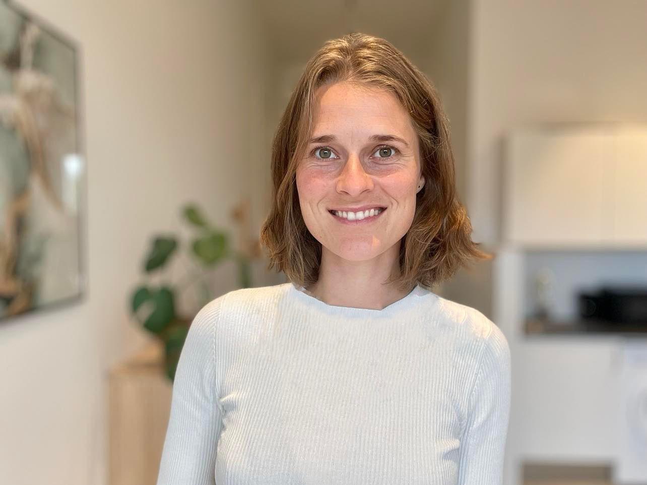 En smilende kvinde med kort hår iført en lys trøje står i et lyst rum med planter i baggrunden.