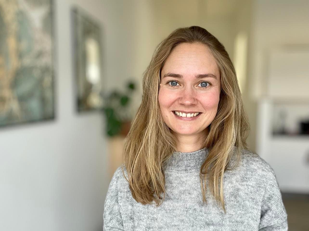En smilende kvinde med langt, lyst hår iført en grå sweater står i et lyst rum med planter i baggrunden.