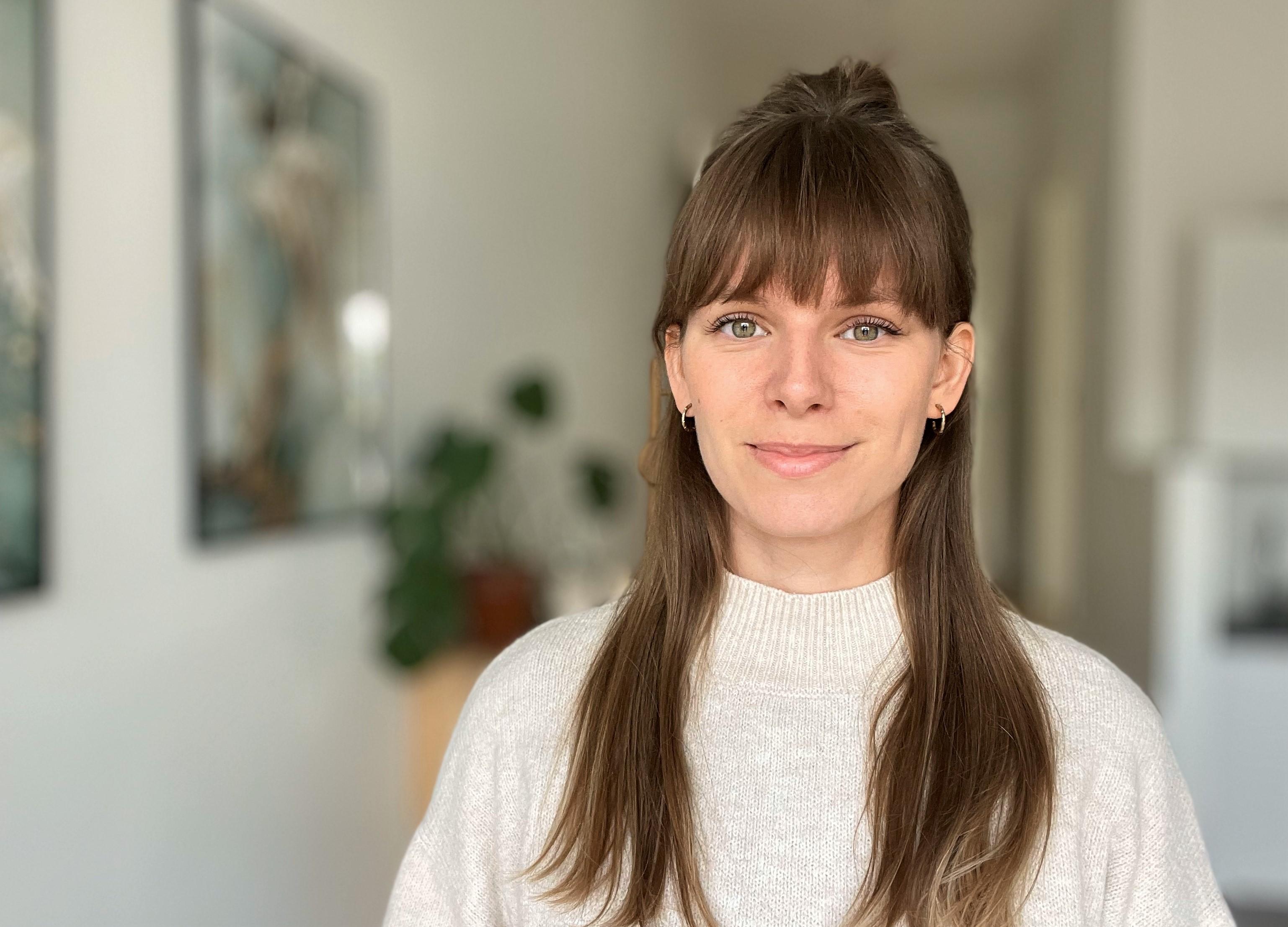 En kvinde med langt, brunt hår iført en lys sweater står i et lyst rum med planter i baggrunden.