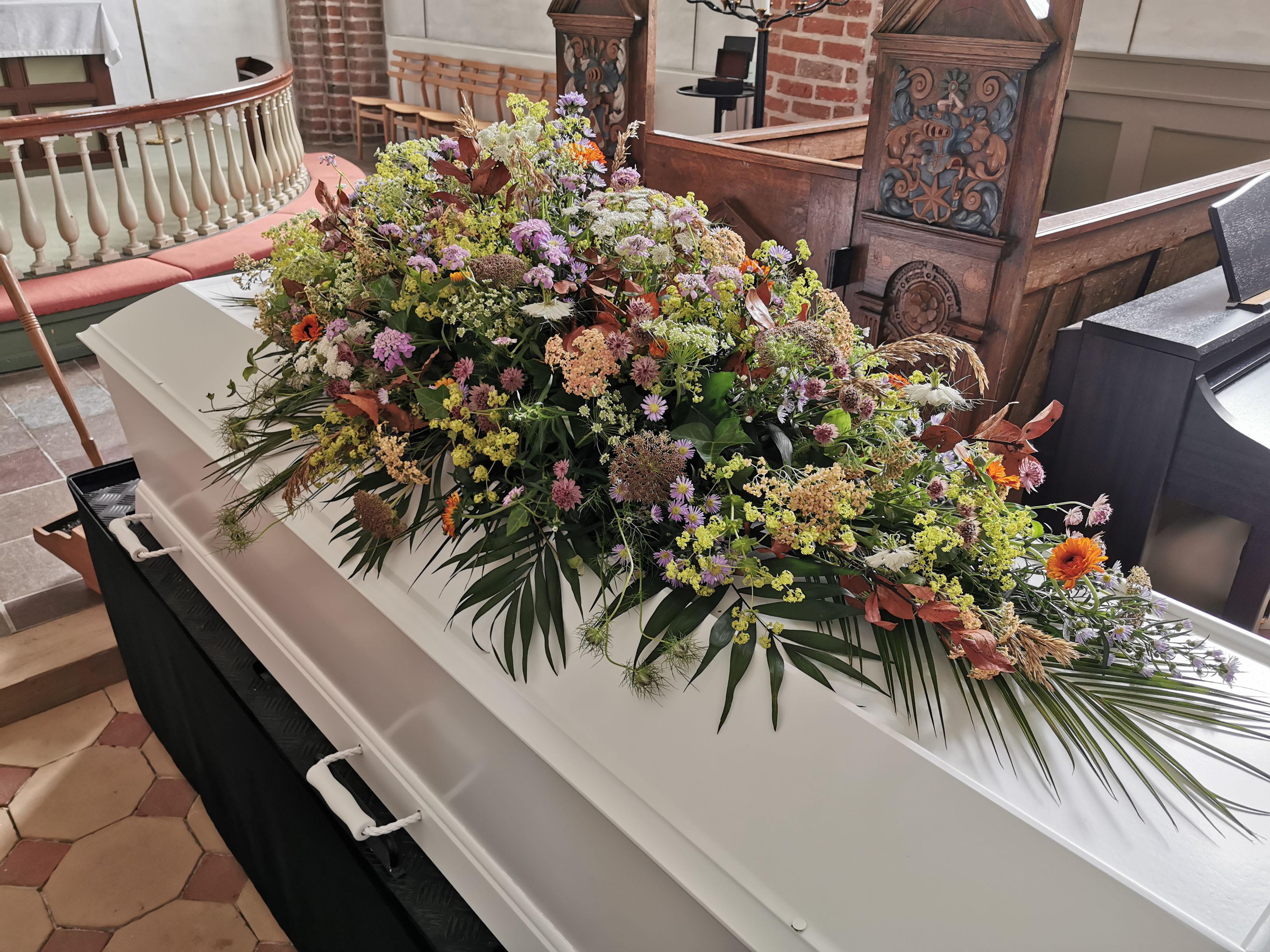 En hvid kiste dekoreret med farverige blomster og grønne planter i en kirke.