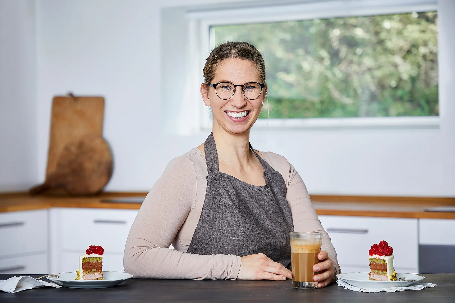 En smilende kvinde i en grå forklæde sidder ved et bord med to kager og en kop kaffe i et lyst køkken.