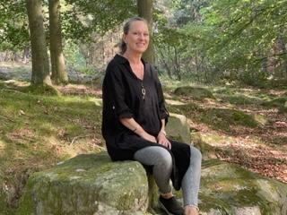 Kvinde i sort kjole sidder på en sten i en skov med grønne træer og mos.