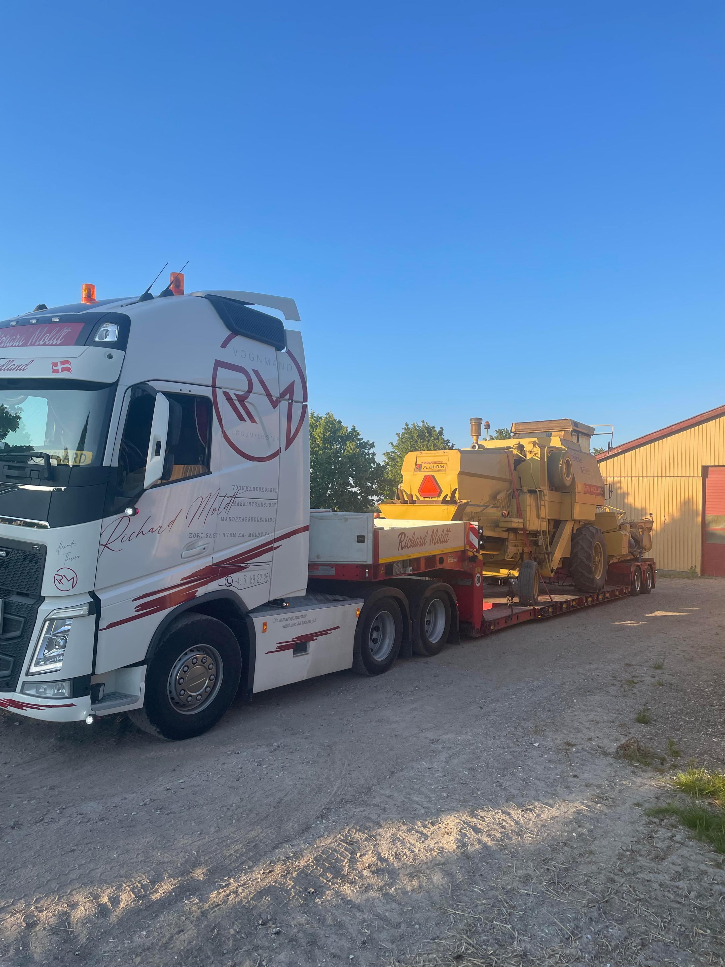 En stor lastbil med en gult maskine på en trailer, parkeret ved en bygning under en klar blå himmel.