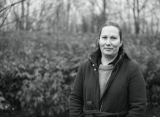 Kvinde i sort-hvid portræt, iført en jakke, står foran en grøn busk i en skovagtig baggrund.