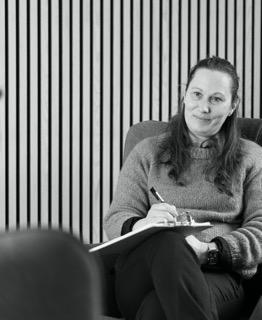 En kvinde med langt hår sidder i en stol og skriver i en notesblok, omgivet af en stribet væg.