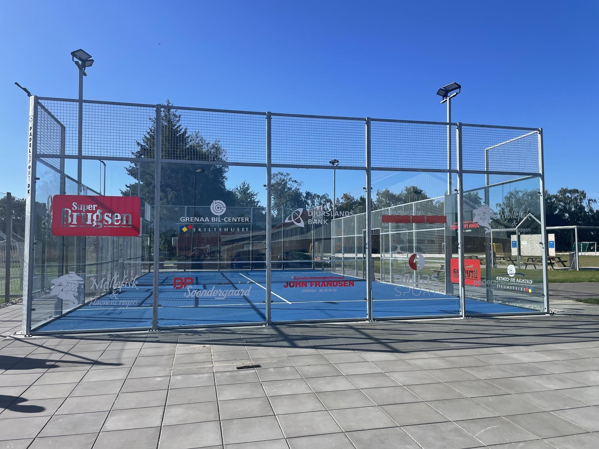 En padelbane med blå belægning og net, omgivet af glasvægge, under en klar blå himmel.