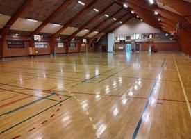 Indendørs sportshal med glat trægulv, højt loft og basketballkurve langs væggene.