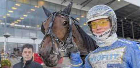 En jockey i blå uniform holder en hest ved siden af en person i baggrunden, med en moderne bygning i baggrunden.