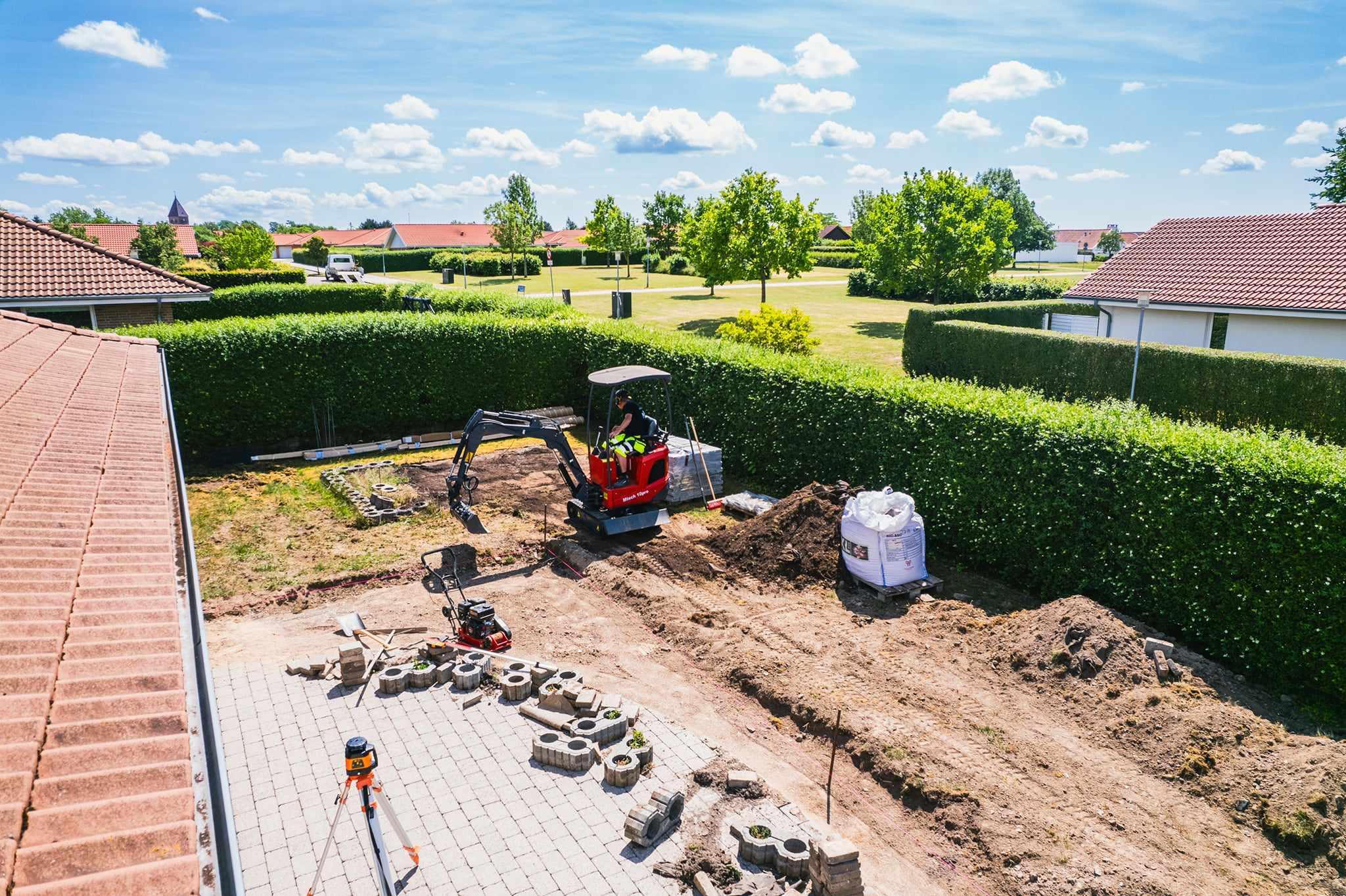 Udsigt over en have med en gravemaskine, der arbejder på en byggeplads omgivet af grønne hække og græsplæner.