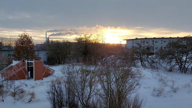 Vinterlandskab med sne, solopgang og røg fra en fabrik i baggrunden.