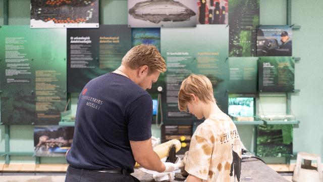 En voksen og et barn ser på en fisk i et udstillingsområde med information om fiskearter.