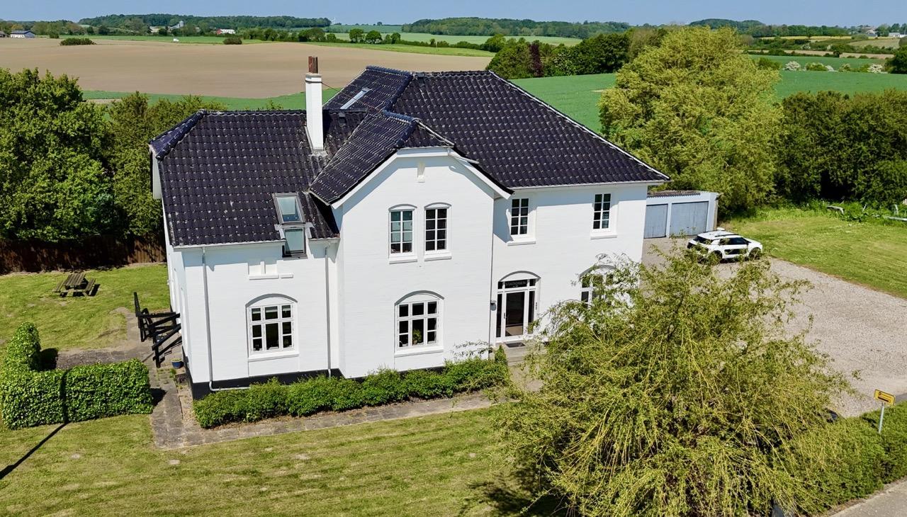 Et stort, hvidt hus med sort tag, omgivet af grønne marker og træer, set fra luften.