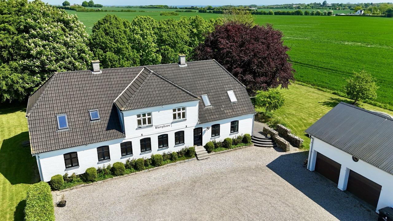 Smukt hus med grå tag og hvid facade, omgivet af grønne marker og træer, med en grusvej foran.