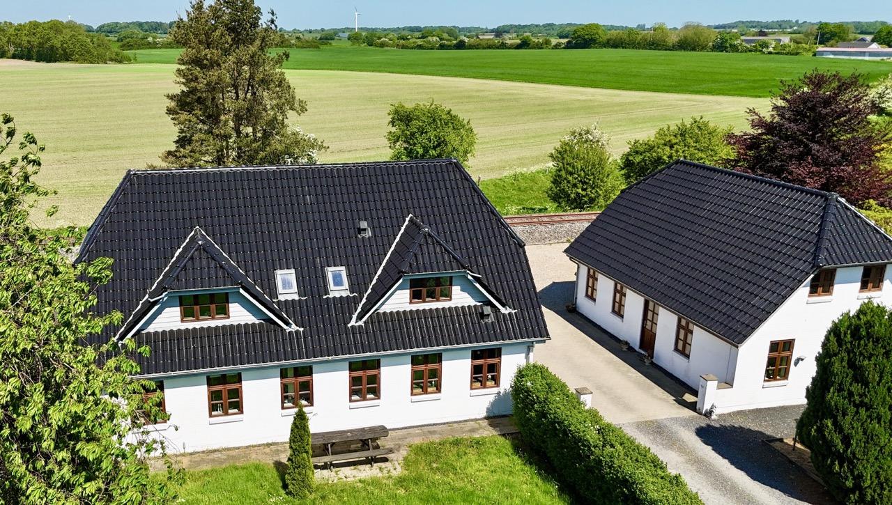 To hus med sort tag og grønne marker i baggrunden, omgivet af træer og en grusvej.