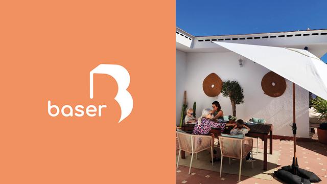 En solrig terrasse med en kvinde, der sidder ved et bord, omgivet af planter og parasoller. Baser-logoet i venstre hjørne.