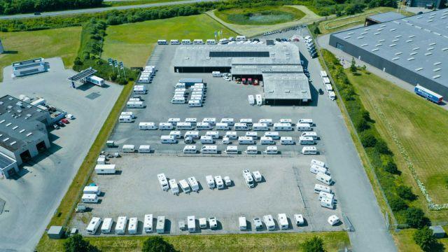 Luftfoto af et stort parkeringsområde med mange campingvogne og en bygning i midten. Grønt område i baggrunden.