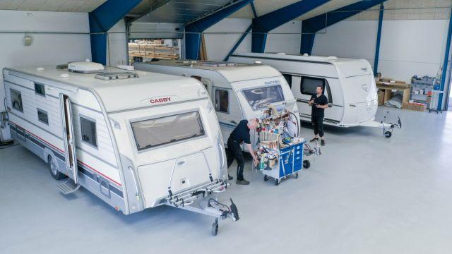 To personer arbejder på campingvogne i en garage med blå bjælker og lys gulv.