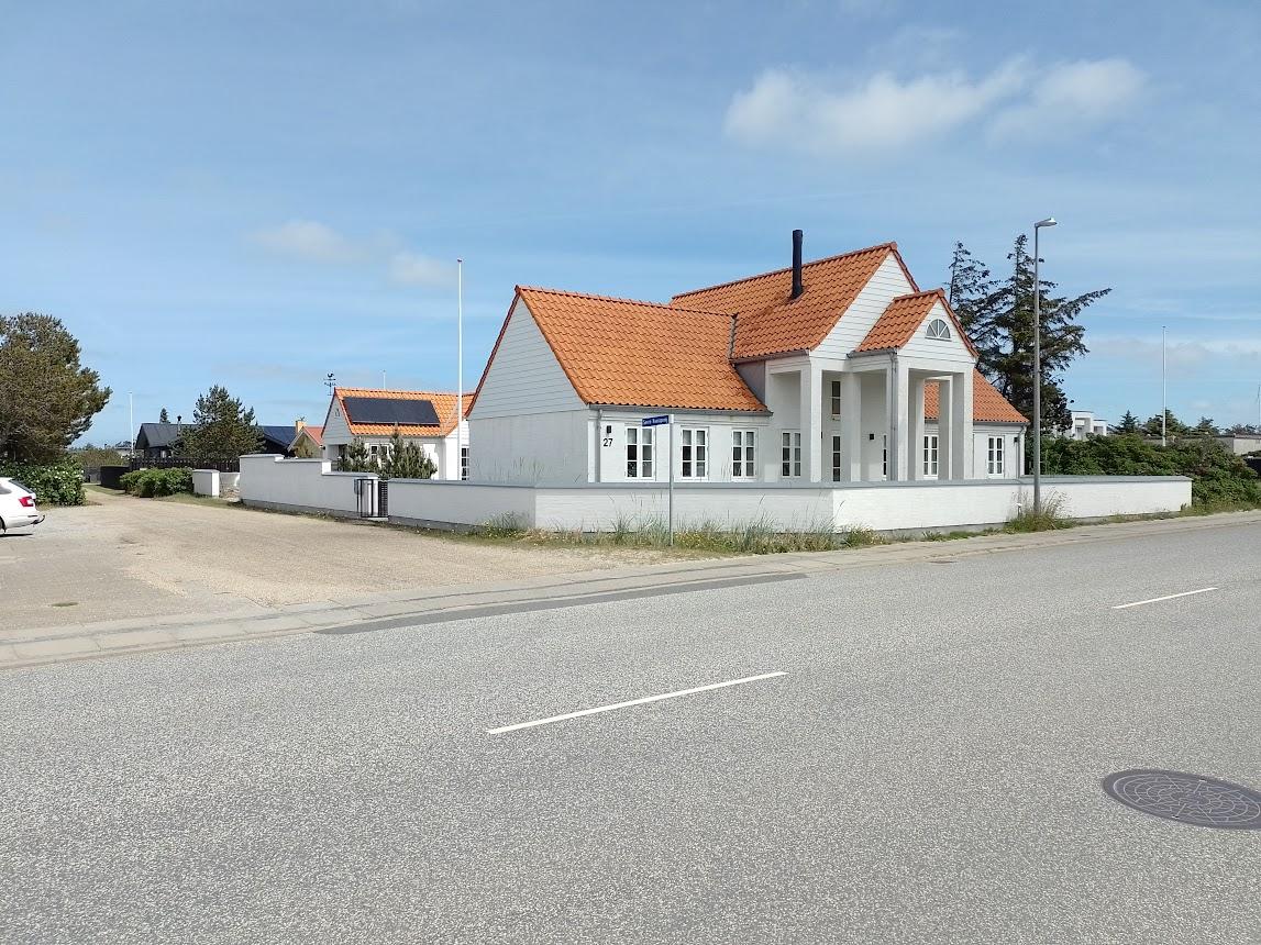 Et hvidt hus med orange tag beliggende ved en vej, omgivet af træer og parkeringspladser.