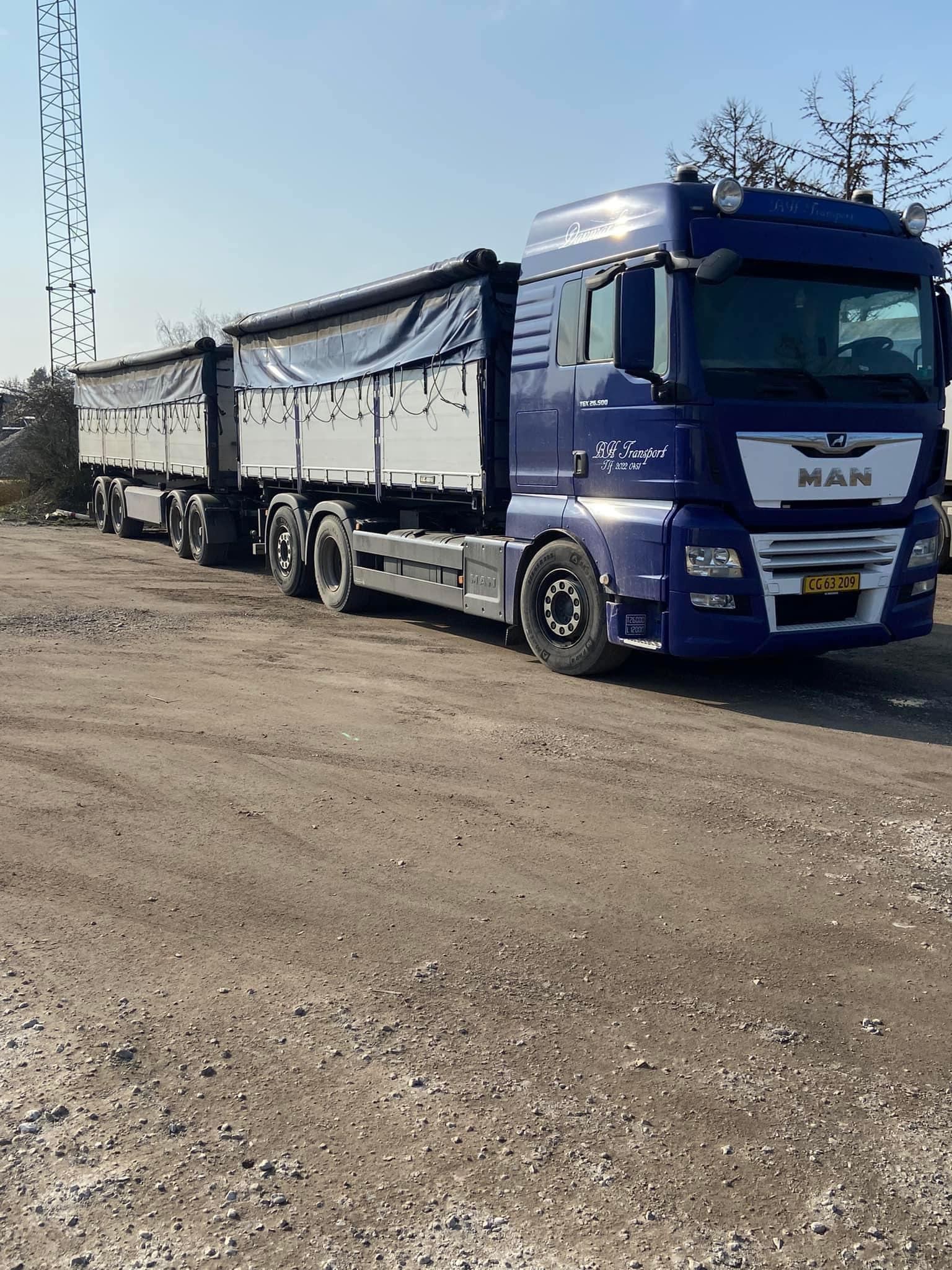 En blå MAN lastbil med en stor trailer parkeret på en grusvej i et åbent område.