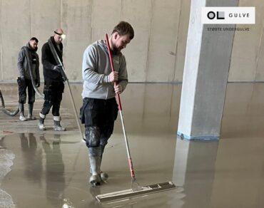 Tre mænd arbejder med at hælde beton på en gulvflade i en stor bygning.