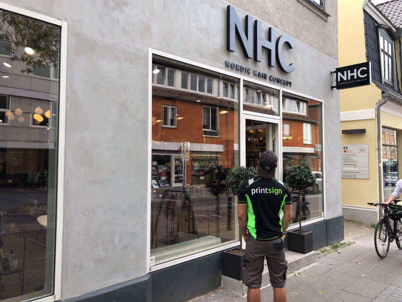 En mand i en sort og grøn trøje står foran en butik med skiltet "NHC" og vinduer, der viser indretningen.